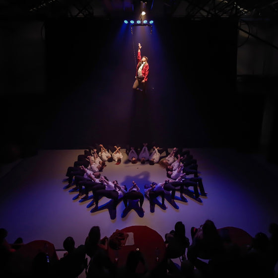 A circle of performers lie on stage, a man is lifted in air with a harness